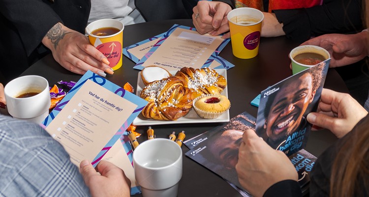 Kanelbullar, mazarin och syltkaka upplagt mitt på runt bord, några personers händer syns på bordet och material om Finansförbundet